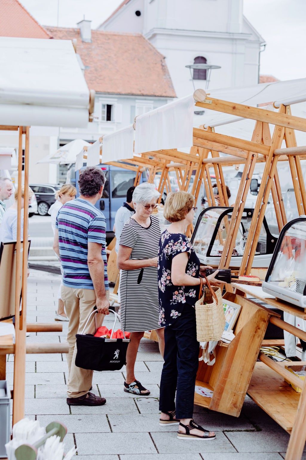 A group of people are shopping at an outdoor market with wooden stalls. One woman holds a basket and another looks at items in a glass case.