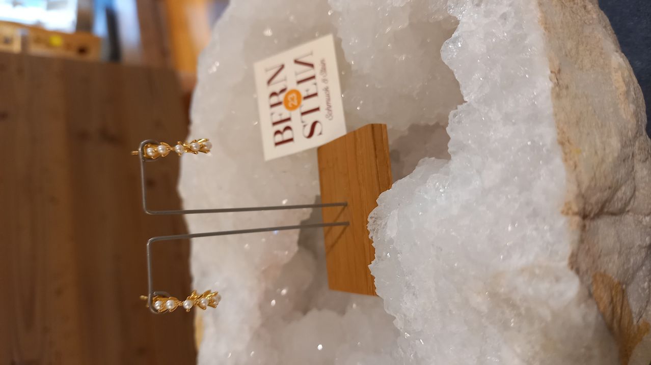 Ein Paar goldene und perlenbesetzte Ohrringe liegen auf einem Holzständer, mit einem weißen Karton, auf dem BERN 2 STEIN steht, und einer kristallartigen Struktur im Hintergrund.