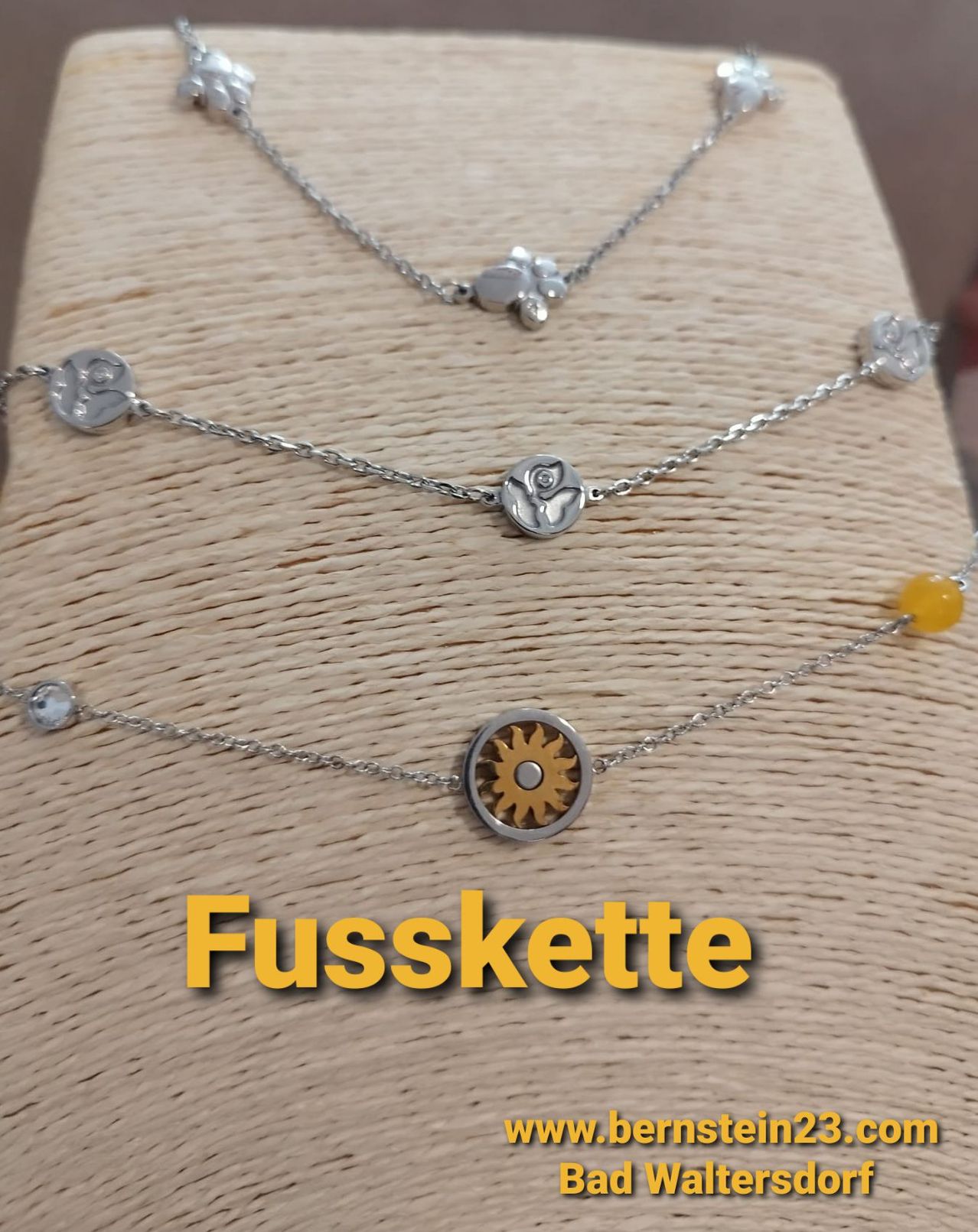 Three silver necklaces on a brown background with a yellow word Fusskette, featuring various pendants like gears and flowers, and a yellow gemstone on the bottom necklace.