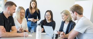 Eine Gruppe von Menschen sitzt bei einer Besprechung um einen Tisch, zwei Frauen lächeln. Ein Laptop und Stifte sind auf dem Tisch, mit Wassergläsern und einer Flasche.