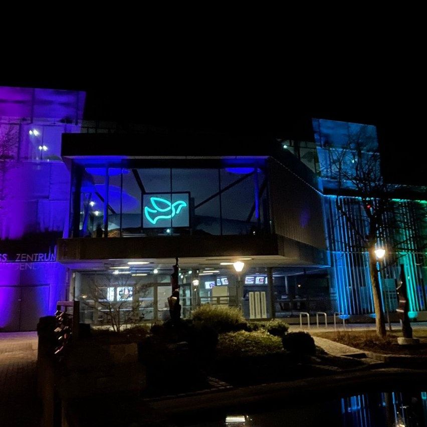 In der Nacht ist das Kongresszentrum mit leuchtenden Regenbogenfarben beleuchtet, mit einem prominenten blauen Taubenlogo und beleuchteten Lampen.