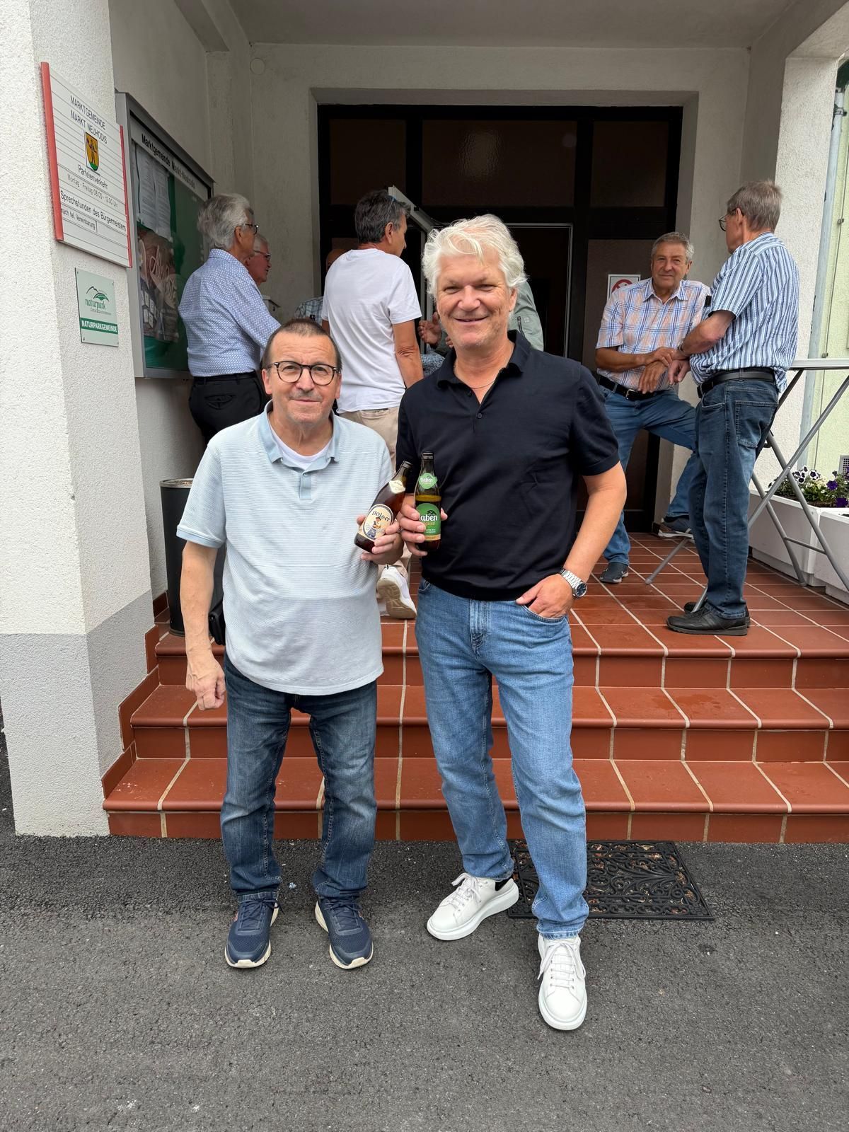 Zwei Männer stehen auf einer Treppe mit Bierflaschen und lächeln. Eine Gruppe von Männern im Hintergrund, einer sitzt auf einem Stuhl.