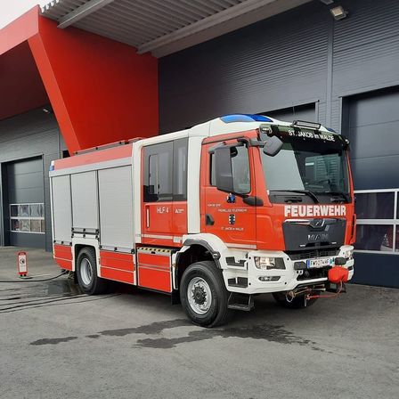 Ein orange-weißer Feuerwehrwagen steht vor einem Feuerwehrhaus mit dem Namen St. Jakob im Walde. Er ist an einen Schlauch angeschlossen und hat eine rote Düse an der Vorderseite.