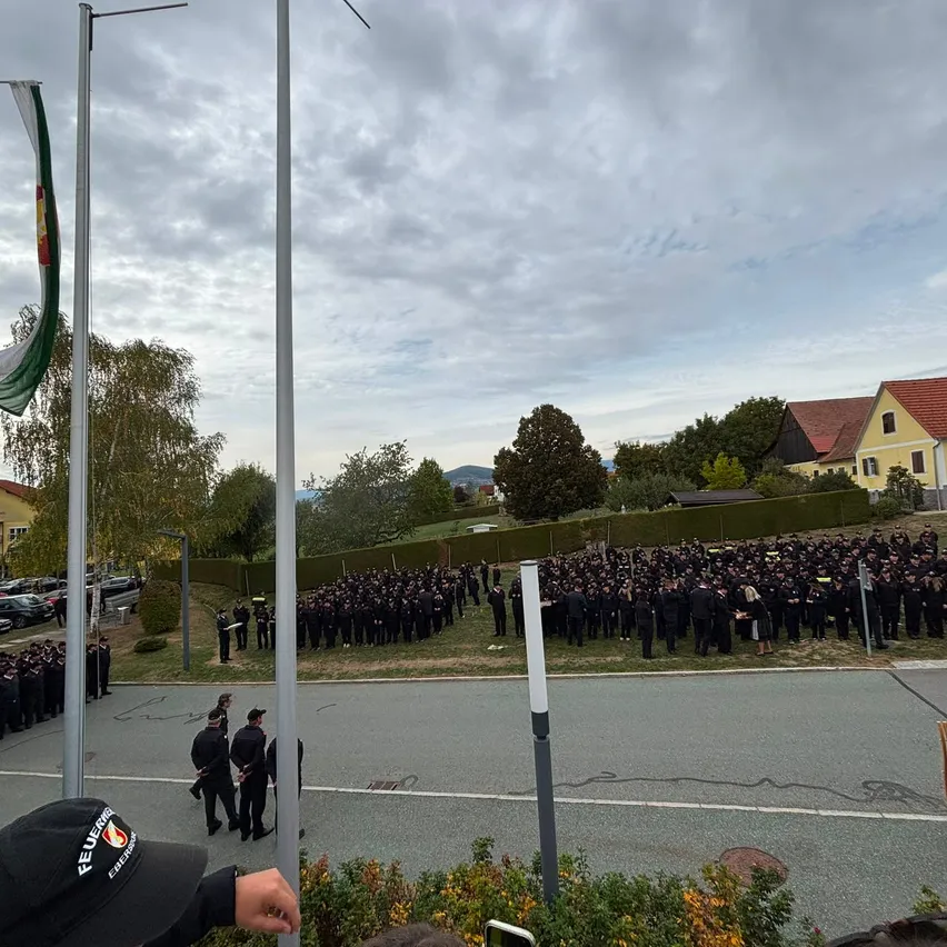 Eine große Gruppe von Menschen in Uniform, möglicherweise Polizeibeamte, ist in einem Außenbereich versammelt. Sie stehen in Reihen, und einige halten Fahnen. Das Gebiet ist von Gebäuden, Bäumen und geparkten Autos umgeben.