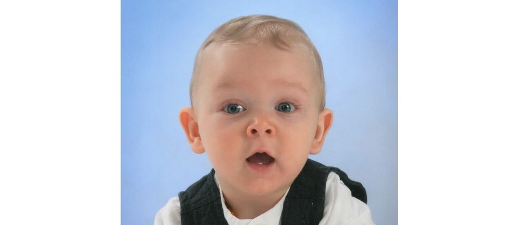 Ein junger Junge mit hellem Haar und blauen Augen, gekleidet in einem weißen Hemd und schwarzem Weste, scheint überrascht vor einem blauen Hintergrund zu sein.