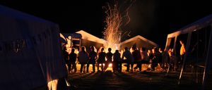 Eine Gruppe von Menschen sitzt nachts um ein Lagerfeuer, mit Zelten und Bäumen im Hintergrund.