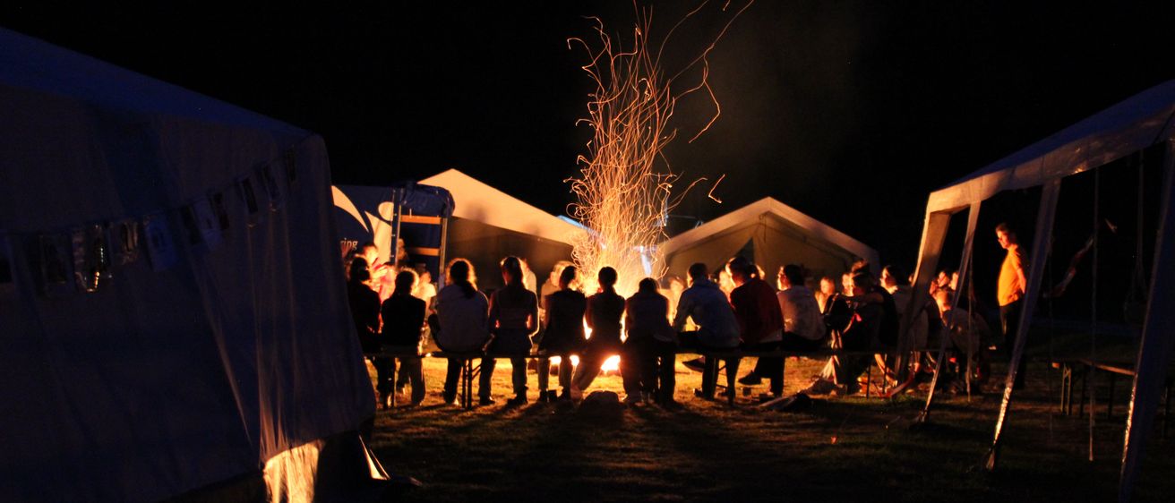 Eine Gruppe von Menschen sitzt nachts um ein Lagerfeuer, mit Zelten und Bäumen im Hintergrund.