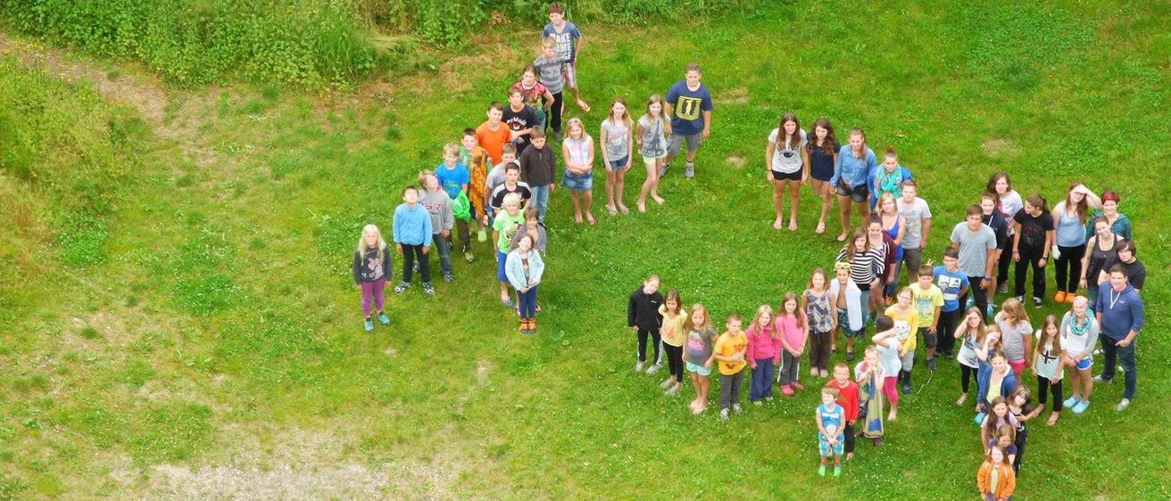 Eine Luftaufnahme einer Gruppe von Kindern, die auf einem Rasenfeld einen Buchstaben 'U' bilden. Sie tragen Freizeitkleidung und Turnschuhe. Links ist ein Steinkreis zu sehen.