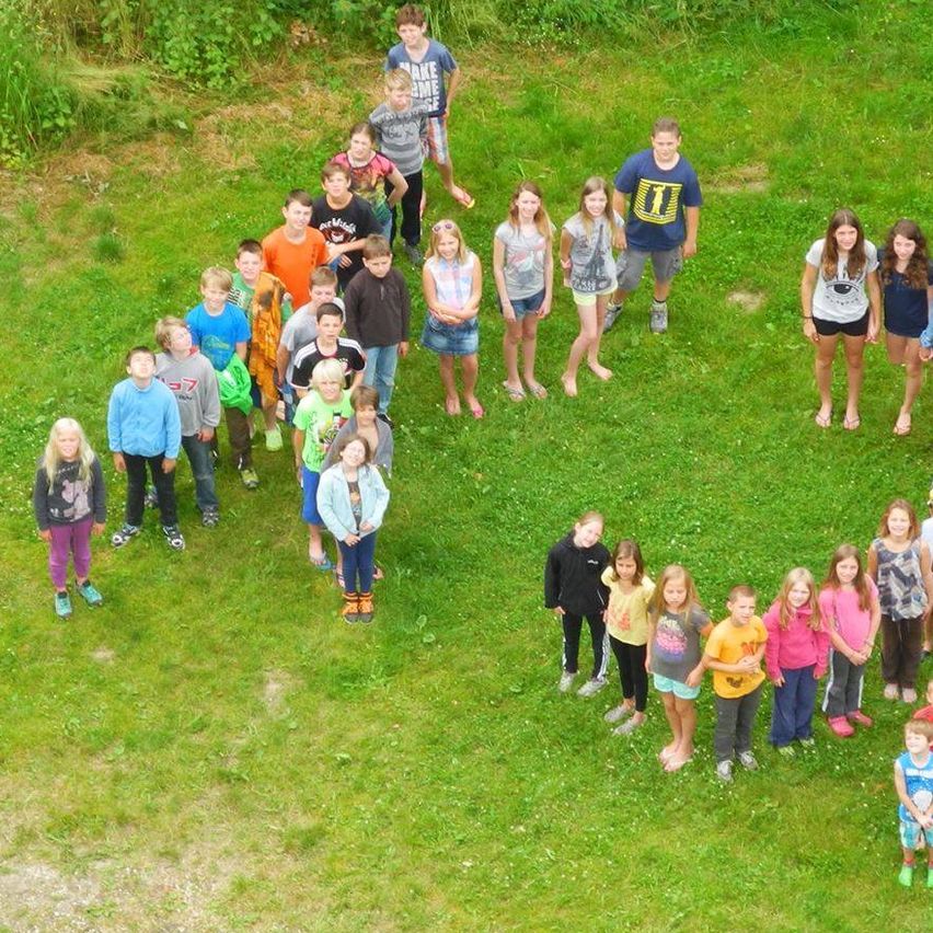 Eine Luftaufnahme einer Gruppe von Kindern, die auf einem Rasenfeld einen Buchstaben 'U' bilden. Sie tragen Freizeitkleidung und Turnschuhe. Links ist ein Steinkreis zu sehen.