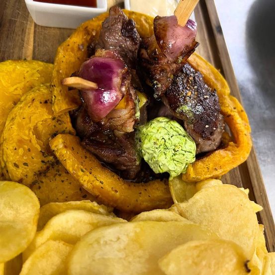 Ein Teller mit gegrillten Fleischspießen auf einem Süßkartoffel-Brötchen mit Avocado, begleitet von Kartoffelchips und einer kleinen Dip-Sauce.