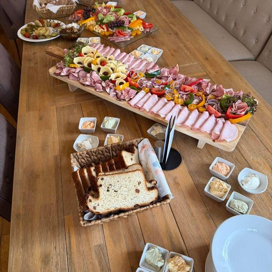 Ein Holztisch zeigt eine große Charcuterie-Platte mit Fleisch, Gemüse und Brot. Verschiedene Dips und Saucen befinden sich in kleinen Schüsseln um die Platte. Teller und Brotkörbe sind ebenfalls auf dem Tisch.