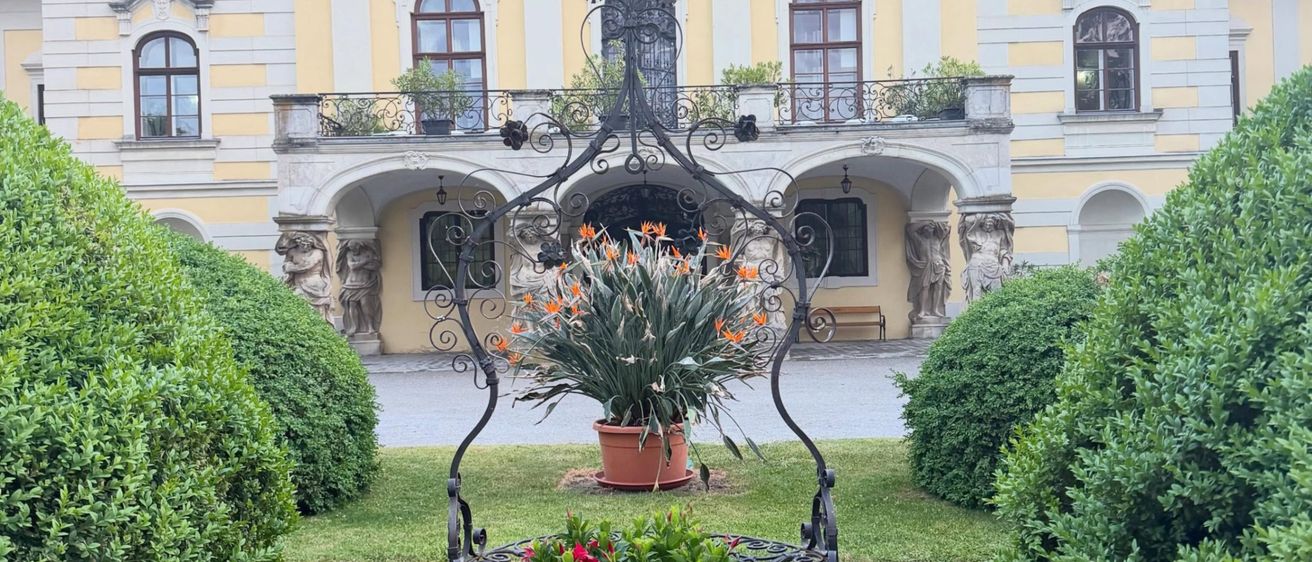 Ein gelbes Herrenhaus mit weißer Einfassung und mehreren Rundbogenfenstern. Davor steht auf einem Rasen ein schwarzer Eisenpflanzenhalter mit Blumen.