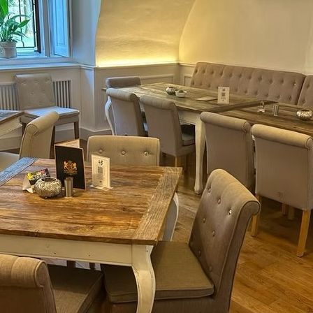 Ein Esszimmer mit mehreren Tischen und Stühlen. Ein Tisch im Vordergrund hat eine Menükarte und Dekorationsartikel. An der Wand befinden sich Topfpflanzen und Heizkörper.