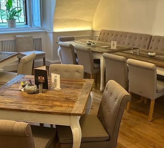 Ein Esszimmer mit mehreren Tischen und Stühlen. Ein Tisch im Vordergrund hat eine Menükarte und Dekorationsartikel. An der Wand befinden sich Topfpflanzen und Heizkörper.