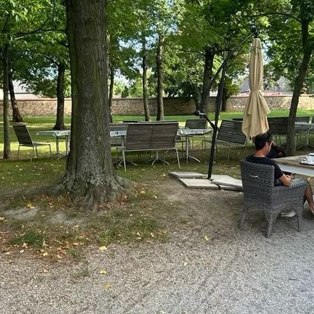 Ein Restaurant im Park hat Tische und Stühle unter Schirmen. Ein Mann und eine Frau sitzen an einem Tisch, und eine weitere Frau steht in der Nähe. Ein Menübrett steht vor einem Baum.