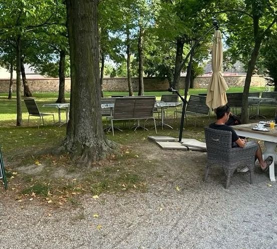 Ein Restaurant im Park hat Tische und Stühle unter Schirmen. Ein Mann und eine Frau sitzen an einem Tisch, und eine weitere Frau steht in der Nähe. Ein Menübrett steht vor einem Baum.