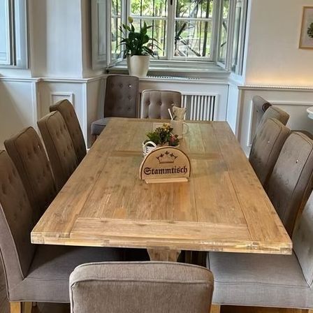 Ein Essbereich mit einem großen Holztisch, Stühlen, einem Topf mit Pflanzen, einer Speisekarte und gerahmter Kunst. Der Tisch hat ein Schild, das Stammtisch steht.