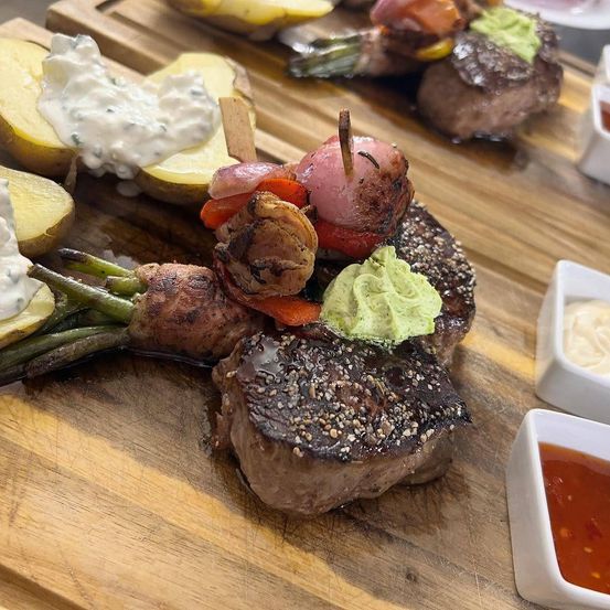 Ein Holzschneidebrett zeigt mehrere gegrillte Steaks, überzogen mit Gemüse, Saucen und Garnierungen. Kleine Schälchen mit Saucen und eine Kartoffel mit weißer Dip umgeben die Steaks.