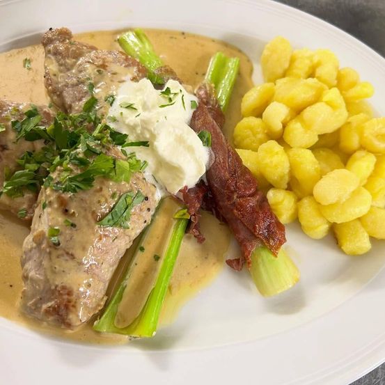 Ein Teller mit zwei Fleischstücken, überzogen mit einer cremigen Sauce, garniert mit Petersilie, begleitet von Spargel, einer Scheibe Prosciutto und Kartoffelpüree.