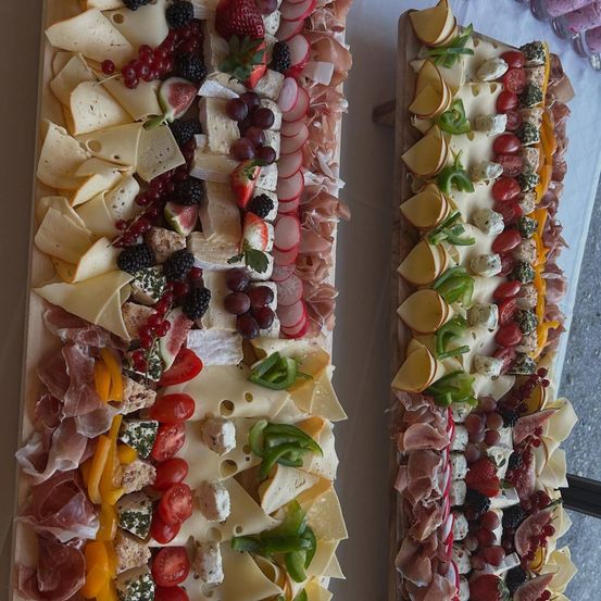 Zwei Bretter mit einer Auswahl an Käse, Fleisch, Früchten und Gemüse ansprechend angeordnet. Die Käsesorten sind geschnitten, und es gibt Trauben, Beeren und geschnittene Paprikaschoten.