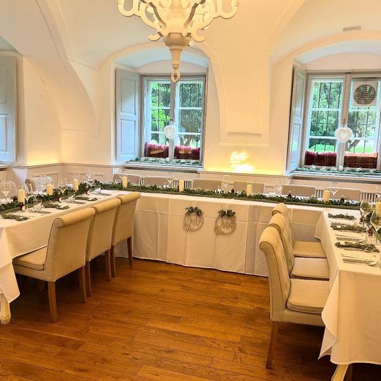 Ein gut dekorierter Speisesaal mit zwei langen Tischen, die für eine Mahlzeit vorbereitet sind. Es gibt weiße Tischdecken, Kerzen und Grünpflanzen auf den Tischen. Der Raum hat Holzböden und große Fenster.