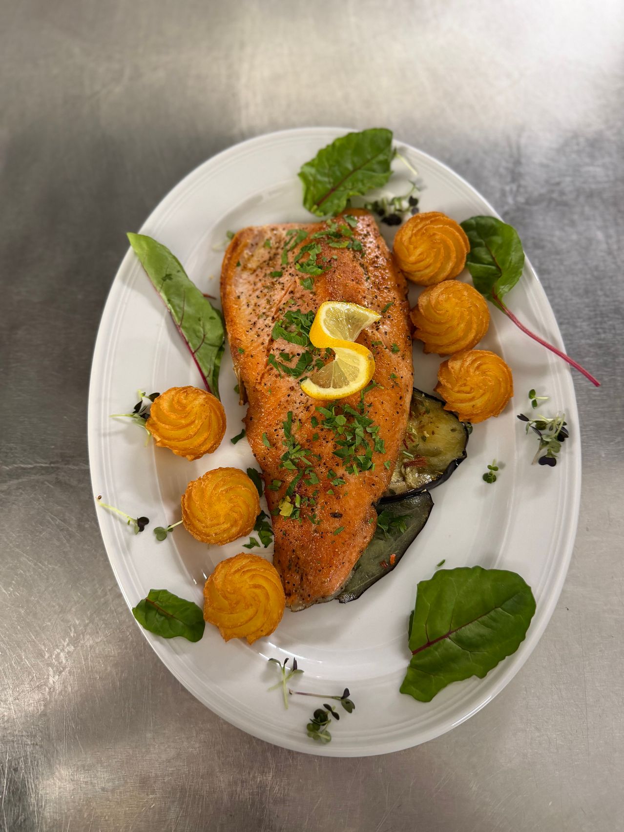 Ein weißer Teller mit gegrilltem Lachs, garniert mit Kräutern und Zitronenscheiben, begleitet von kleinen gebackenen Kartoffelkegeln und grünen Blättern.