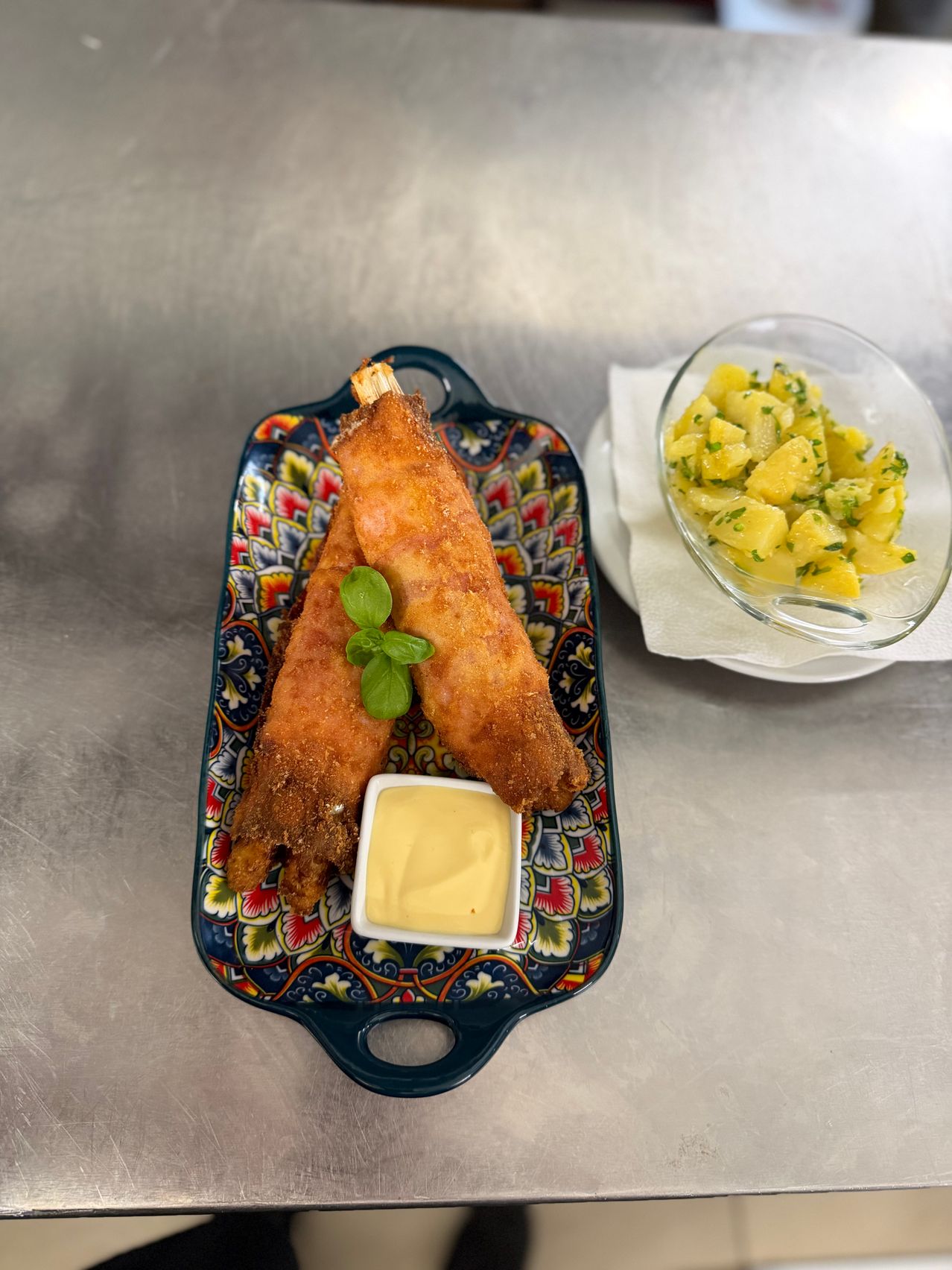 Zwei frittierte Lebensmittel mit Sauce, begleitet von einer Schüssel Kartoffelsalat auf einem Tablett. Der Hintergrund ist eine saubere, metallische Oberfläche.