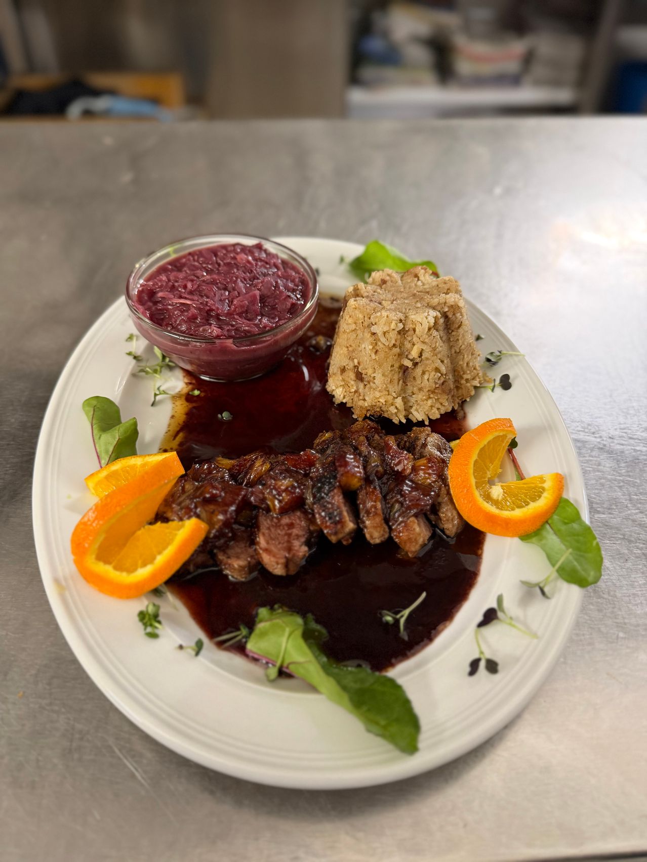 Ein Teller mit gegrilltem Fleisch in Sauce, Orangenscheiben, Rotkohl und Dessert. Das Fleisch und die Sauce haben eine reiche, dunkle Farbe, während die Orangenscheiben leuchtend sind.