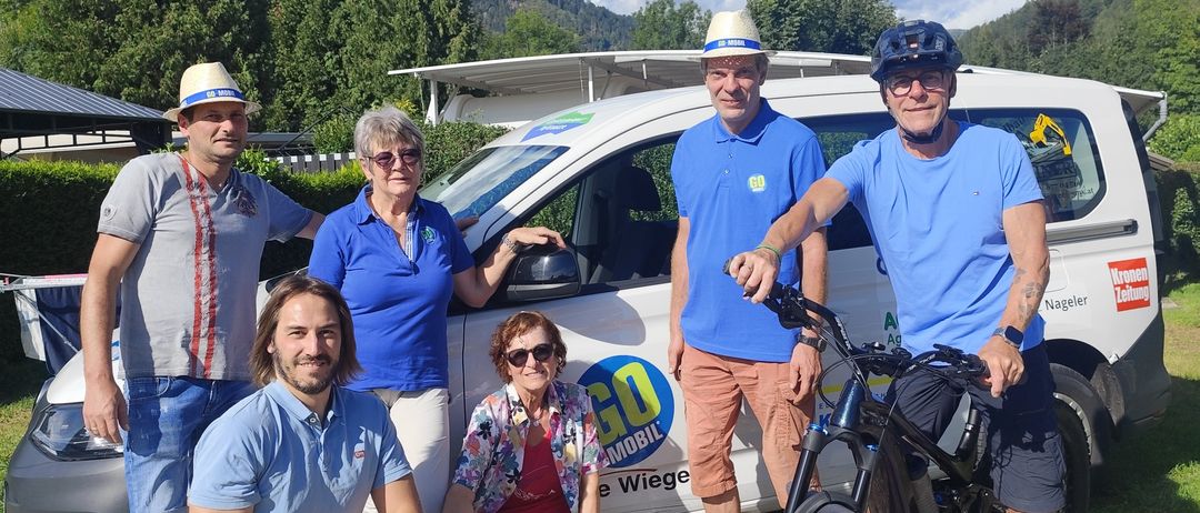 A group of people stand in front of a van with a bicycle, all wearing hats and sunglasses. The van has a logo and text on it. They are on a gravel road with mountains in the background.
