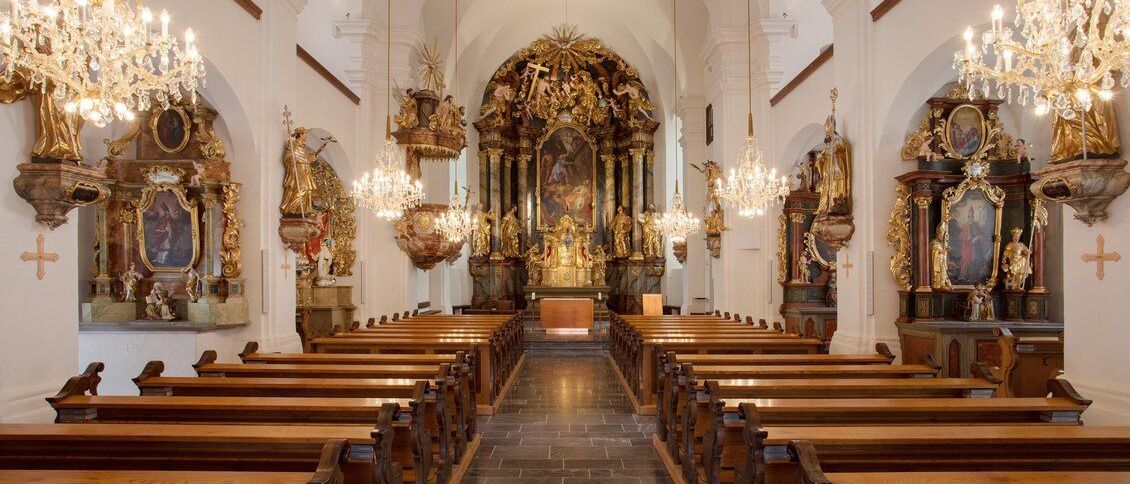 Ein prächtiger Kircheninnenraum mit hölzernen Kirchenbänken, einem goldenen Altar und kunstvollen Schnitzereien. Kronleuchter hängen von der Decke, und Statuen schmücken die Wände.