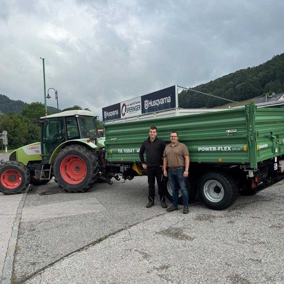 Zwei Männer stehen neben einem grünen Traktor und Anhänger mit einem Banner, das #Husqvarna und Sprenger auf einem bewölkten Tag liest.