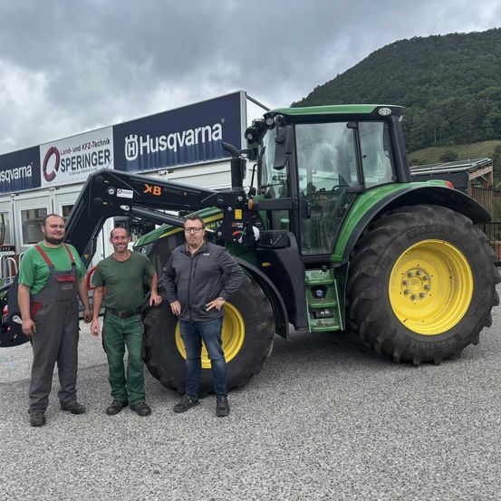 Drei Männer stehen vor einem großen grünen Traktor mit einem gelben Rad, in der Nähe eines Gebäudes mit einem Schild für Husqvarna.
