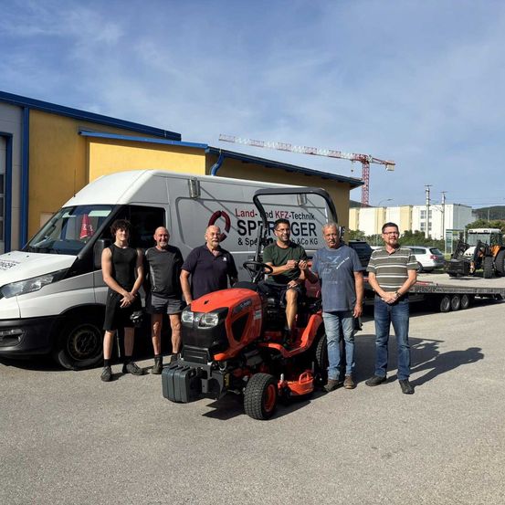 Sechs Männer stehen vor einem weißen Van und einem roten Rasenmäher. Der Van trägt den Schriftzug 'Land-KFZ-Technik Sprenger & Spd & Co KG'. Der Rasenmäher hat den Schriftzug 'Sprenger'. Dahinter steht ein Gebäude mit einem Kran.