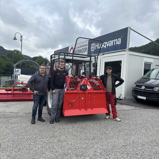 Drei Männer stehen neben einer roten Maschine mit Käfig. Hinter ihnen ist ein Gebäude mit dem Husqvarna-Logo und ein Volkswagen-Van zu sehen.