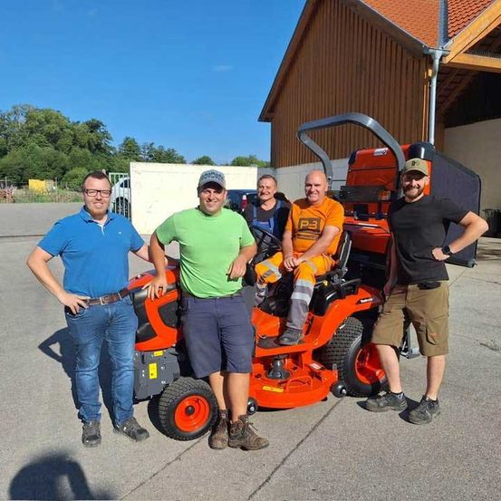 Fünf Männer stehen um einen roten Rasenmäher. Ein Mann sitzt auf dem Rasenmäher. Hinter ihnen ist ein Gebäude mit einer weißen Wand.