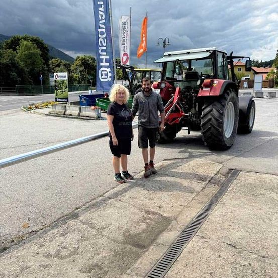 Ein Mann und eine Frau stehen neben einem roten Traktor am Straßenrand, mit Bannern und Pflanzen im Hintergrund.
