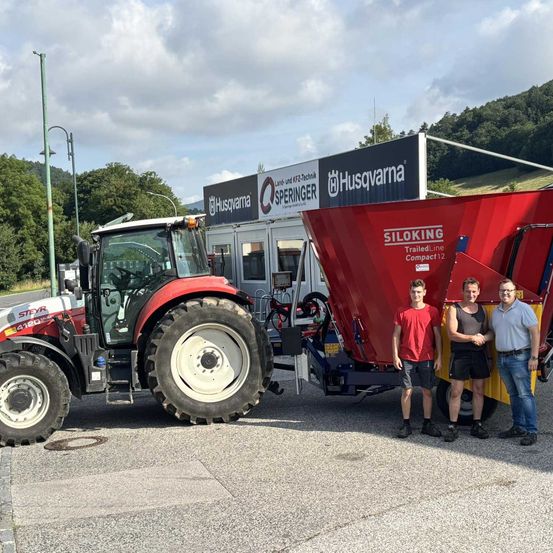 Drei Männer stehen neben einem roten Traktor und einem roten Siloking-Anhänger vor einem Gebäude mit dem Namen Sprenger.
