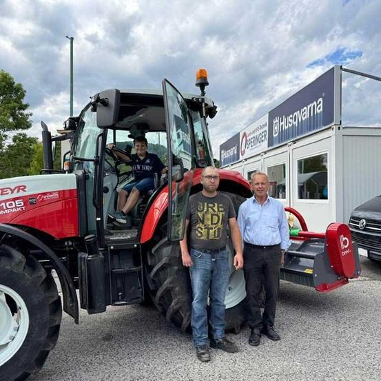 Drei Männer stehen vor einem roten Traktor mit einem Schild, das Husqvarna liest. Der Mann auf der linken Seite trägt eine Brille und Turnschuhe, während der Mann in der Mitte ein graues T-Shirt trägt. Der Mann auf der rechten Seite trägt ein blaues Hemd.
