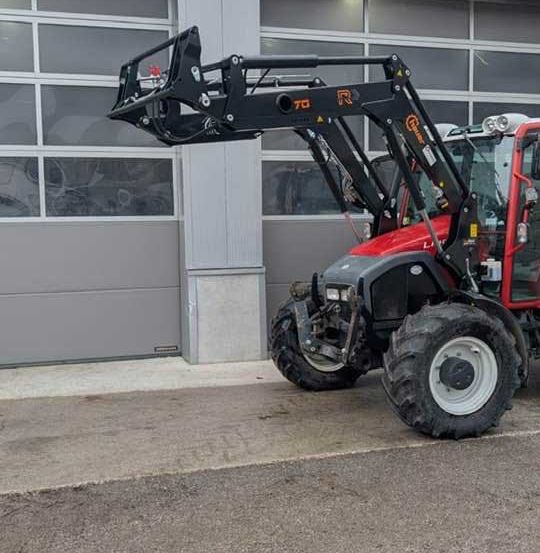 Ein roter Traktor mit schwarzem Anbaugerät steht vor einem Gebäude mit Glastüren.