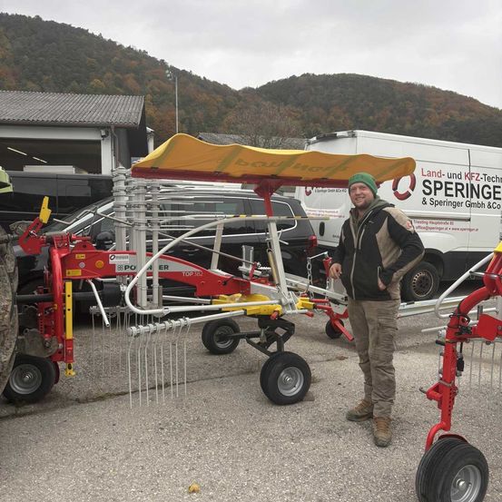 Ein Mann steht neben einem großen landwirtschaftlichen Fahrzeug, einer rot-gelben Maschine mit einem gelben Verdeck. Das Fahrzeug hat große Räder und eine hohe Metallstruktur. Hinter dem Fahrzeug steht ein weißer Van mit dem Text 'Land- und KFZ-Technik Speringer & Speringer GmbH & Co. KG'.