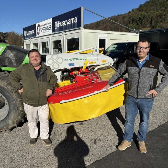 Zwei Männer stehen vor einer rot-gelben Maschine, wahrscheinlich einem Traktor, im Freien. Hinter ihnen befindet sich ein Gebäude mit dem Namen Husqvarna auf einem Schild.