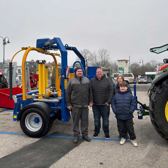 Eine vierköpfige Familie posiert vor einem blauen Traktor mit gelber Anbauvorrichtung. Sie befinden sich auf einem Parkplatz im Freien mit mehreren Autos und einem großen Fahrzeug im Hintergrund.