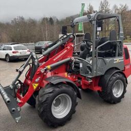Ein roter Wiedemann Traktor mit einem gelben Dreieck an der Front und einem silbernen Pflug steht auf einem Parkplatz neben mehreren Autos.