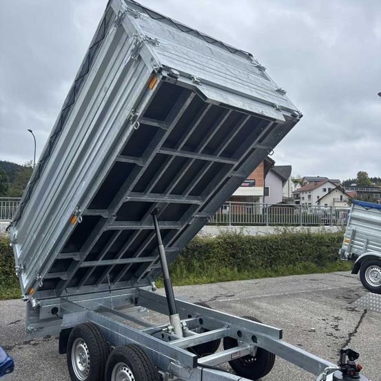 Ein offener Metall-Kippanhänger steht auf einem Schotterplatz, mit Häusern und einer Straßenlaterne im Hintergrund.