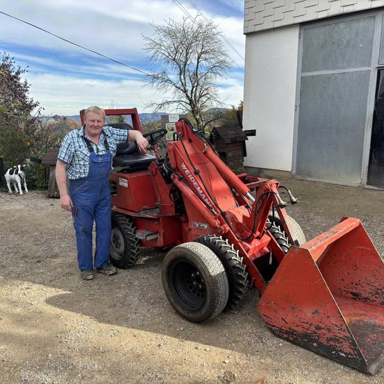 Ein Mann steht neben einem roten Traktor und lehnt sich mit einem Lächeln daran. Hinter ihm steht ein Scheunengebäude mit einer kleinen Hundestatue. Bäume und ein Haus sind im Hintergrund.
