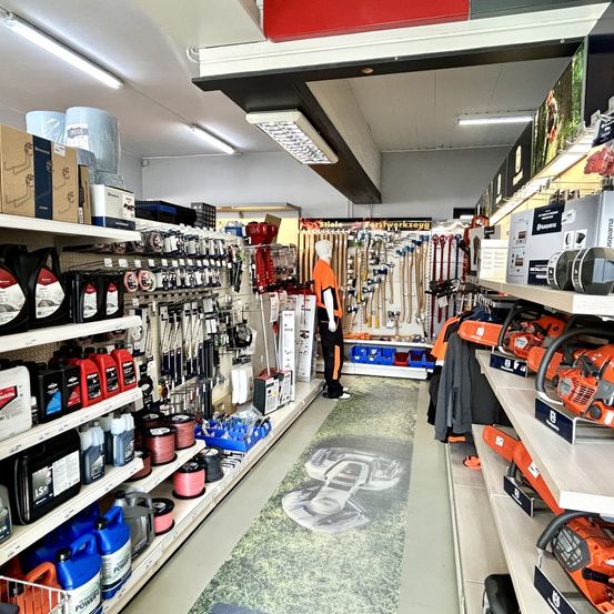 Ein gut organisierter Laden mit verschiedenen Produkten im Angebot, einschließlich Kettensägen, Ölen und anderen Werkzeugen, mit einem Mannequin in einem orangefarbenen Outfit in der Mitte.