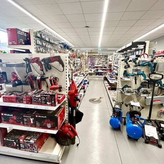 Ein gut beleuchteter Gang in einem Baumarkt mit verschiedenen Werkzeugen und Geräten zur Schau, darunter rote und schwarze Elektrowerkzeuge, Kreissägen und Luftkompressoren.