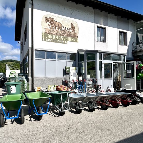 Eine Reihe von Schubkarren und Transportwagen steht vor einem Gebäude mit einem Schild, das 'Landmaschinen Springer' liest. Das Gebäude hat Glasfenster und Türen, und es gibt einige Topfpflanzen und eine Uhr auf dem Balkon.