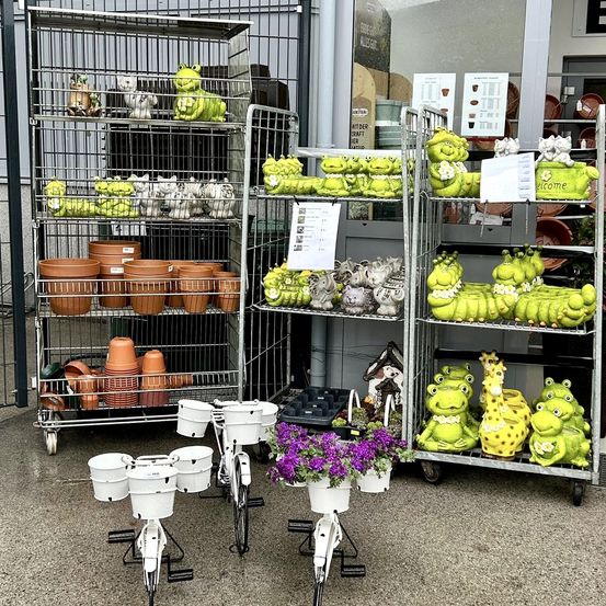 Ein Gartenladen zeigt verschiedene Keramik-Froschfiguren und Töpfe auf Metallregalen. Die Regale sind mit grünen Froskskulpturen und weißen Töpfen gefüllt. Ein weißes Fahrrad mit einem Blumenvase steht vor den Regalen.