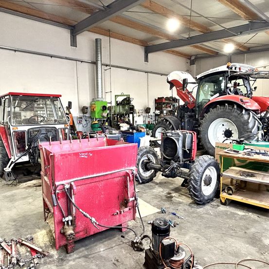 Eine Werkstatt mit mehreren Traktoren und Maschinen, darunter ein roter Traktor mit einer weißen Kabine und einem roten Frontlader.